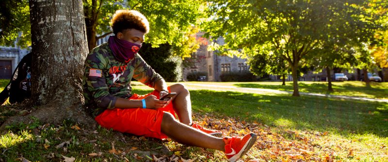 a young man, dressed in Hokie style, reads while sitting on the ground beneath a tree - the afternoon sun shines down on him