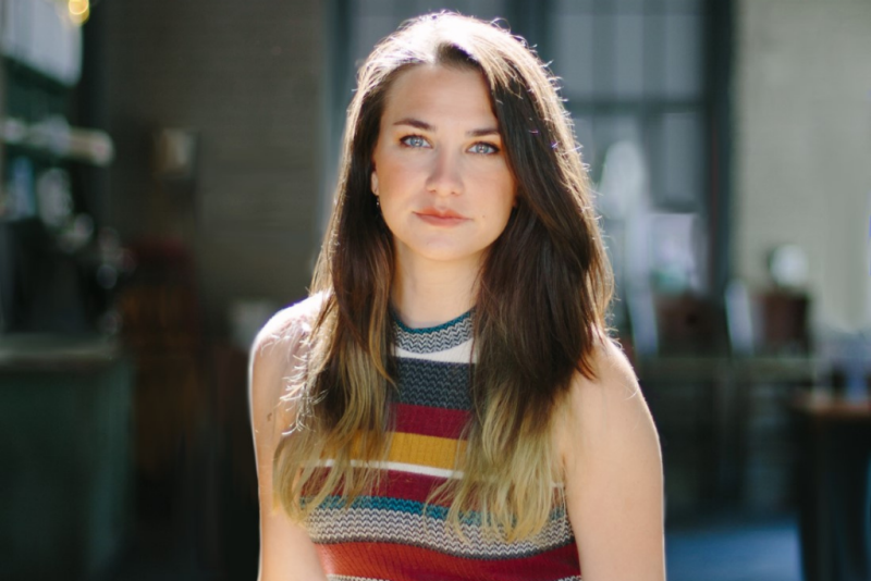 Dr. Bethany Lackey, SOE alumna, confidently looks at the camera. Her long brown hair has blonde tipped ends, and she wears a body conscious striped sleeveless sweater dress