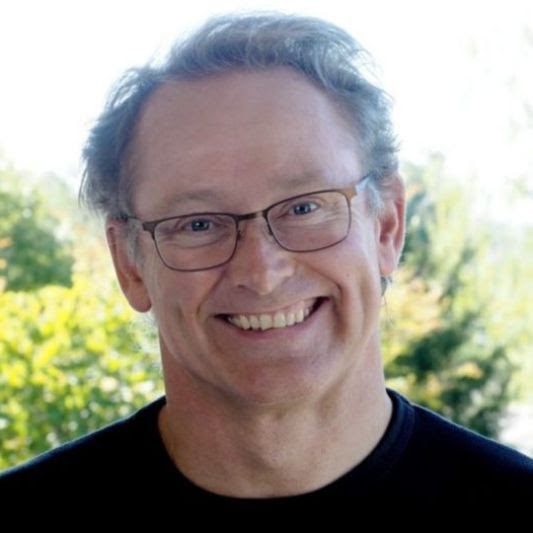 Dr. David Hicks smiles at the camera, wearing a black tshirt, standing in front of some greenery