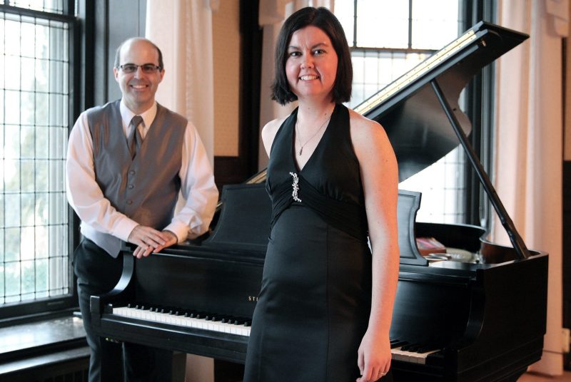 Gregory Millar and Lisa Raposa stand beside a piano.