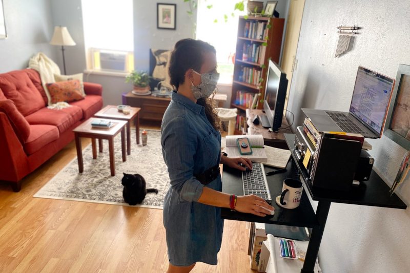 Whitney Hayes stands at a computer workstation in her home. Her cat joins her in the room.
