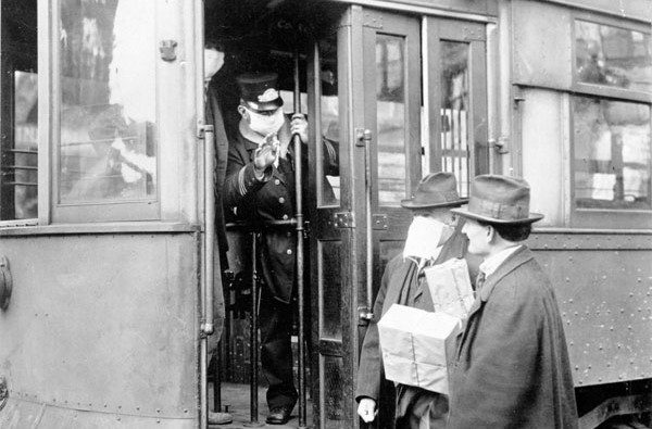 A street car conductor wearing a mask is not allowing passengers aboard without a mask during the 1918 flu pandemic. Courtesy of National Archives. The conductor is leaning out the front door of the trolley to speak to two men.