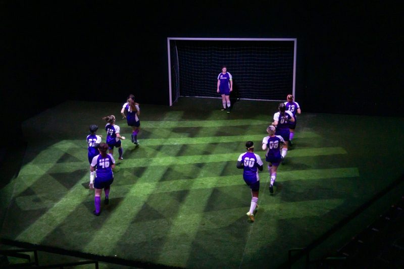 Theatre set featuring soccer players in blue uniforms at the goal line. Green netting is reflected on the ground. 
