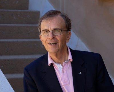 dr alexander leans casually against a stair railing. He is wearing a navy blazer over a lightly unbuttoned pink shirt, and is smiling comfortably