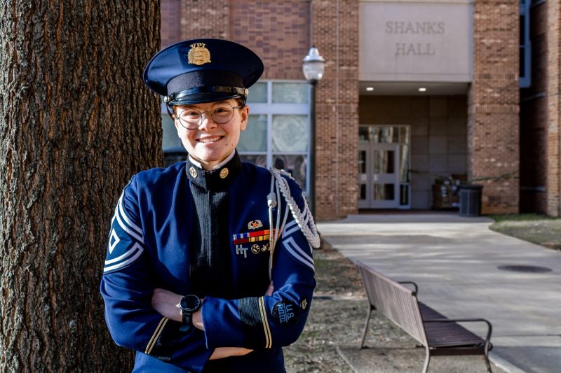 A female member of the corps wears her dress uniforma and smiles at the camera. She stands in front of a building with a sign that reads Shanks Hall.