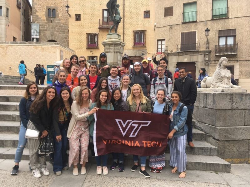A group of students stands in front of a staircase while holding a Virginia Tech flag.