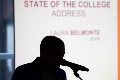 Dean Belmonte speaks at the podium in front of the projection screen. The screen causes only her silhouette to be visible.
