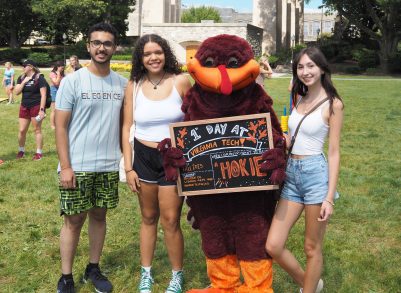 Students pose for a photo with the HokieBird during a sunny day on the Drillfield. Students are smiling while the HokieBird holds a sign that says "1st Day of Classes"