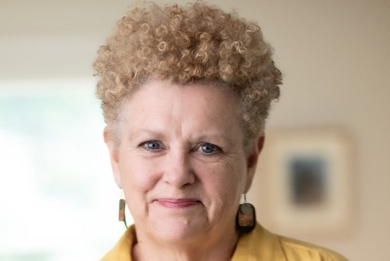 Woman with short blond curly hair, wearing earrings and a yellow blouse.